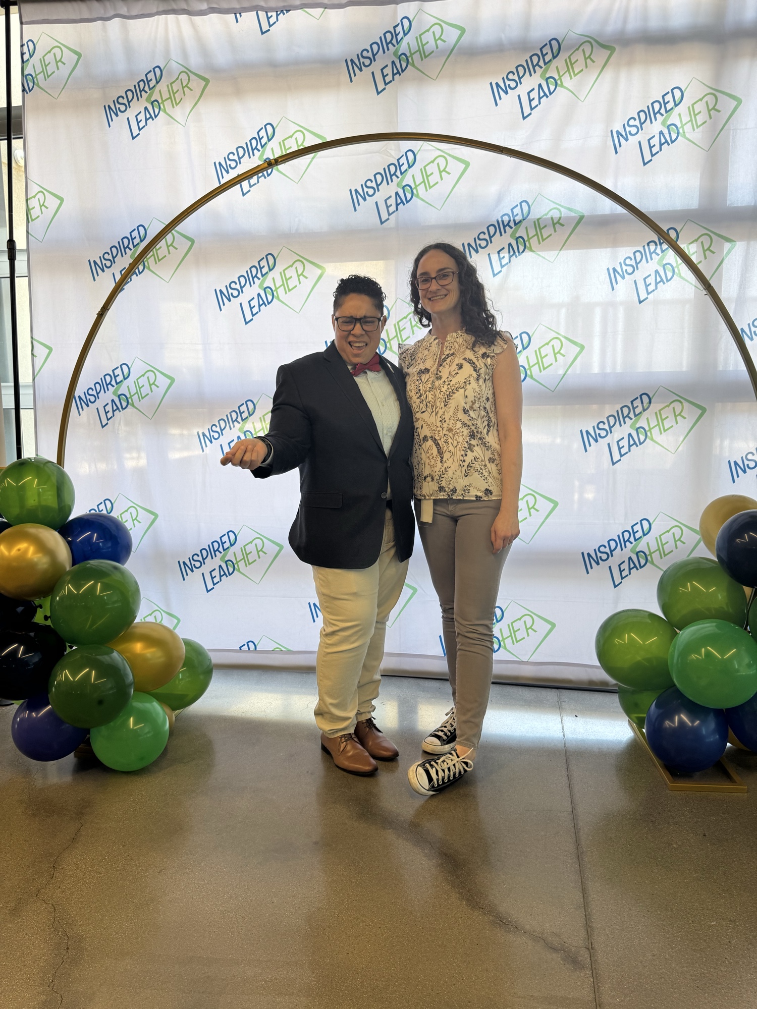 Two Inspired LeadHER 2025 graduates posing under the gold balloon arch with the Inspired LeadHER backdrop on graduation day.
