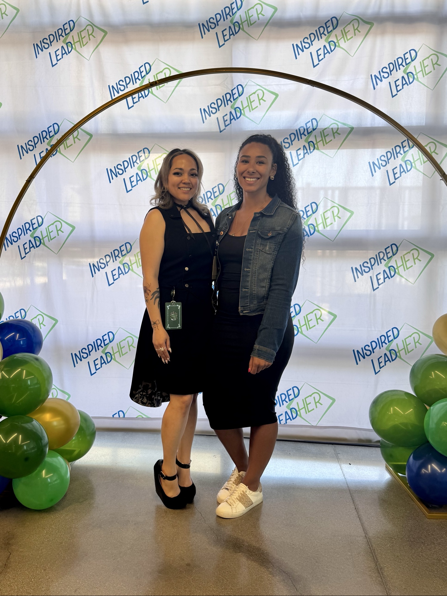 Two Inspired LeadHER 2025 graduates posing together under the gold balloon arch on graduation day.