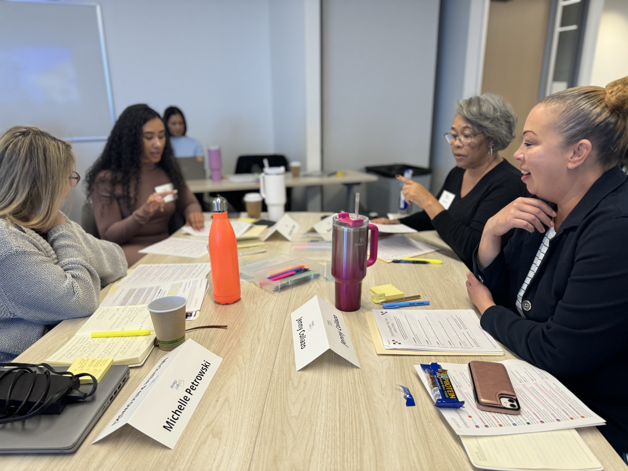 Four women in animated discussion around a wood table with name tents and coffee cups during an Inspired LeadHER 2025 cohort session.