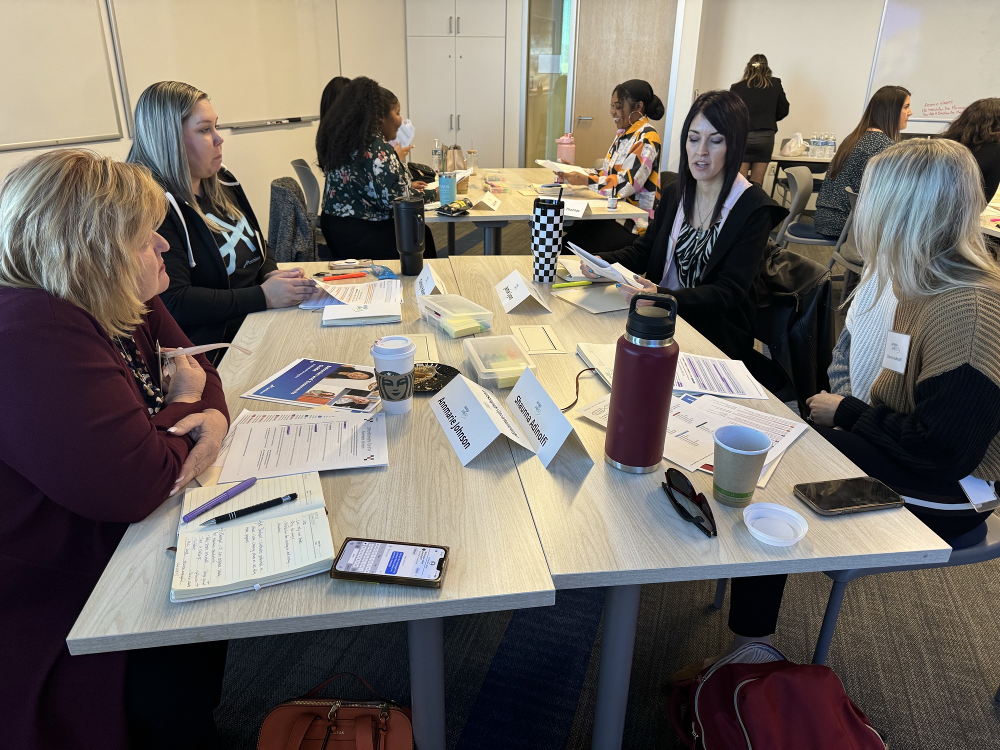 Five women in animated discussion around a round table with name tents, water bottles, and worksheets during a 2025 Inspired LeadHER cohort session at the Achieve campus, Tempe AZ.